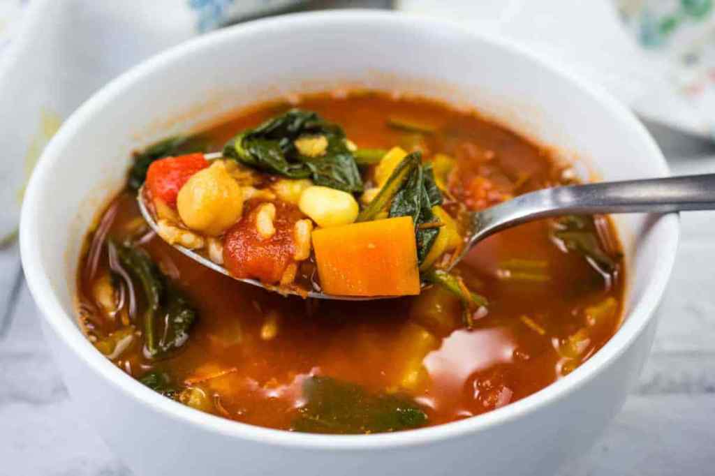 A spoonful of colorful vegetable soup in a white bowl, showcasing ingredients like spinach, peppers, and beans.
