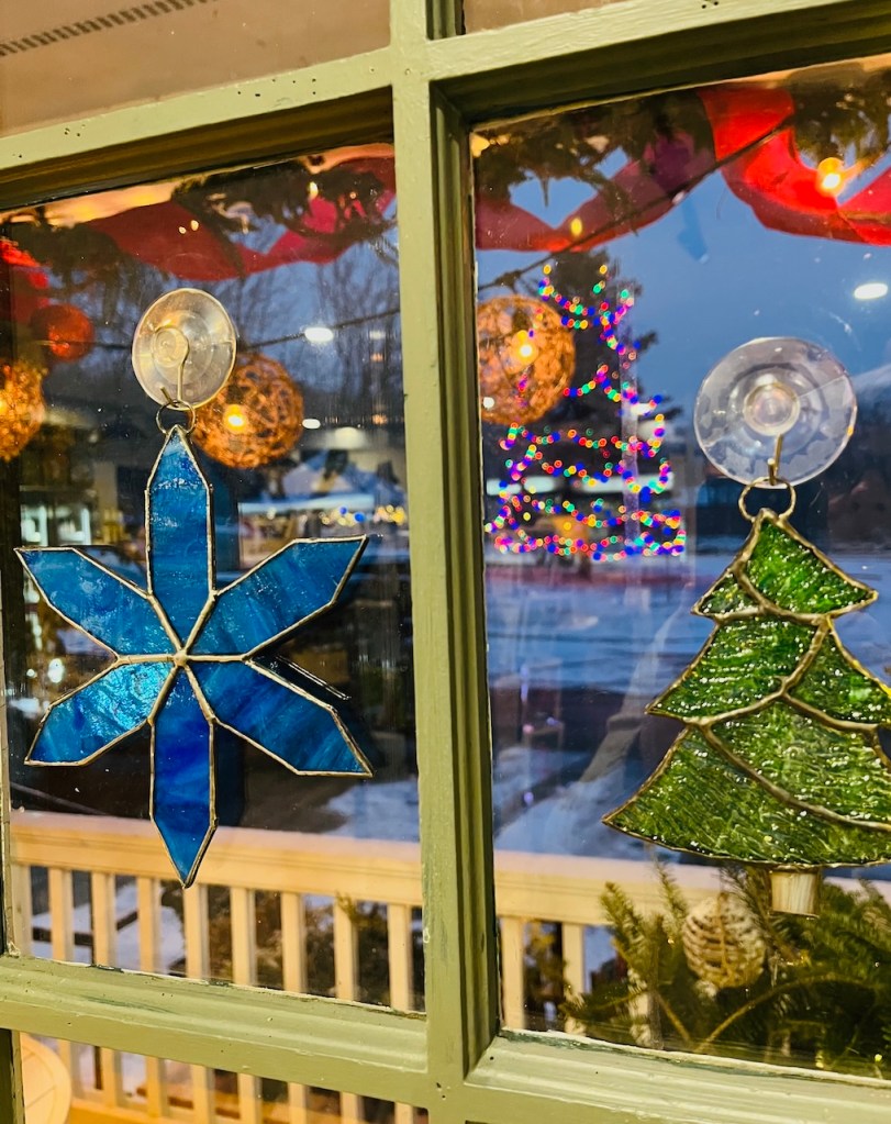 Stained glass decorations in the shape of a snowflake and a Christmas tree hanging in a window, with festive lights visible in the background.