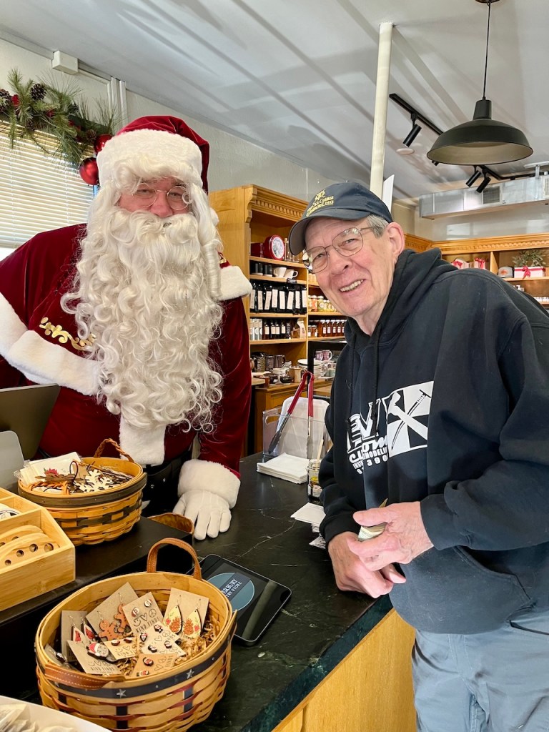 A person dressed as Santa Claus standing behind a counter with a customer, both smiling. The setting features wooden shelves with products and baskets displayed.