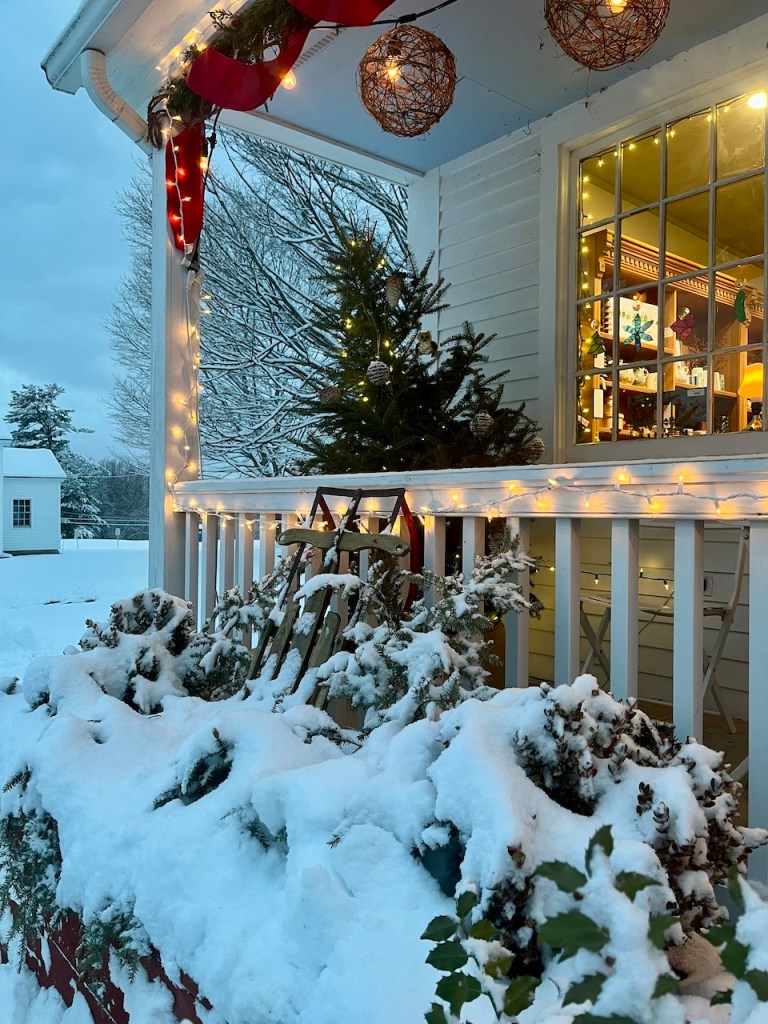 A cozy winter scene featuring a porch decorated for the holidays, with twinkling lights, a small evergreen tree, and snow-covered plants.