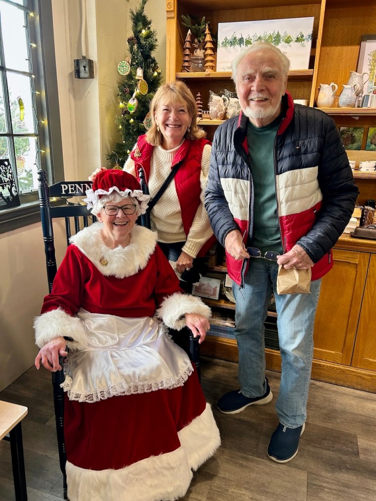 Three people posing together in a cozy store, with a holiday theme. One person is dressed as Mrs. Claus, wearing a red dress with white trim, while the others are casually dressed, smiling and standing beside her.