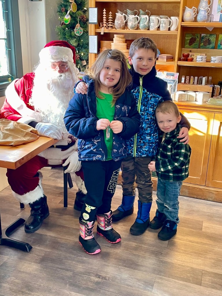 Three children standing in front of Santa Claus in a festive setting, with a decorated background and seasonal decorations.