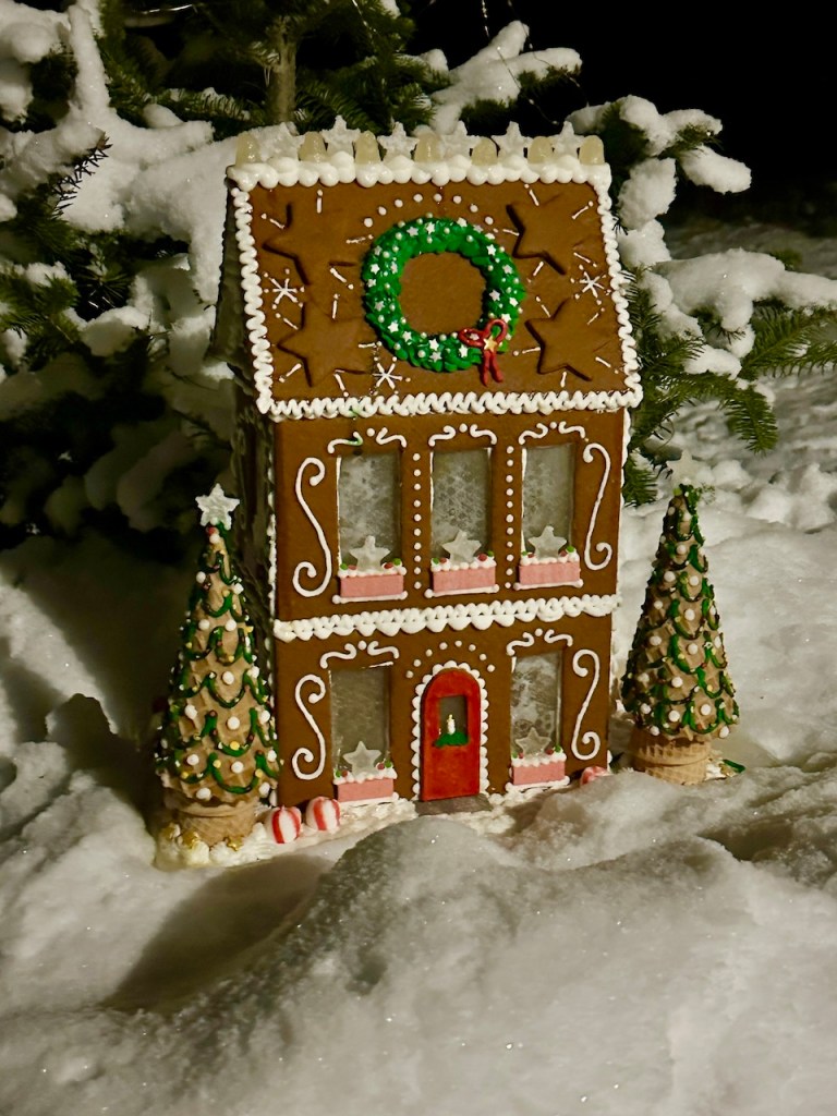 A beautifully decorated gingerbread house surrounded by snow, featuring a green wreath on the roof, candy canes at the base, and two festive trees on either side.
