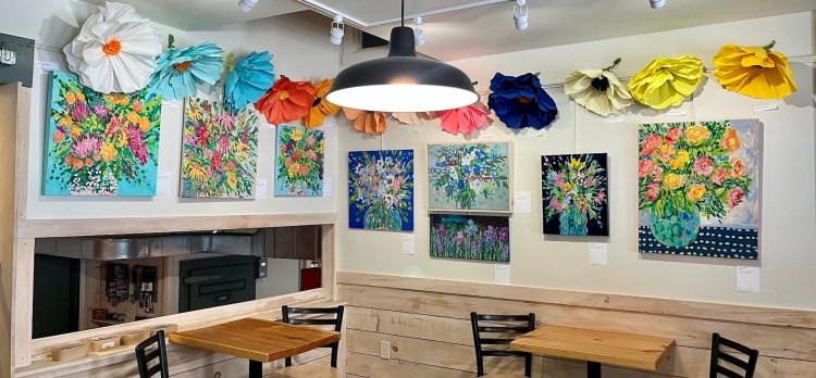 Colorful floral paintings displayed on a café wall, with large paper flowers hanging above them. Two wooden tables with black chairs are in the foreground.