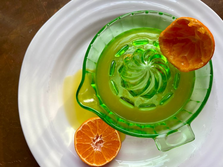 A green glass juicer with a small amount of freshly squeezed orange juice and a halved orange on a white plate.