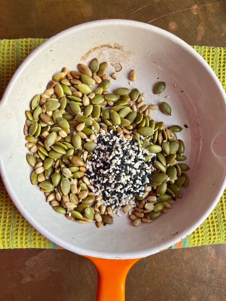 A white frying pan filled with a mix of green pumpkin seeds, sunflower seeds, and sesame seeds, placed on a textured green cloth.