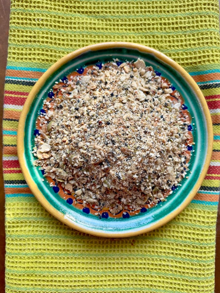 A colorful ceramic plate filled with a coarse mixture of ground spices and grains, placed on a textured green fabric with colorful stripes.