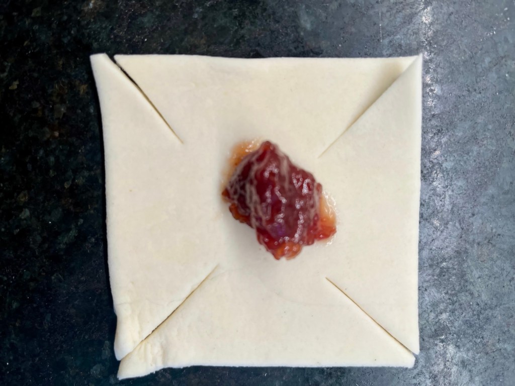 A square pastry dough with folded corners, topped with a dollop of red fruit jam, placed on a dark surface.