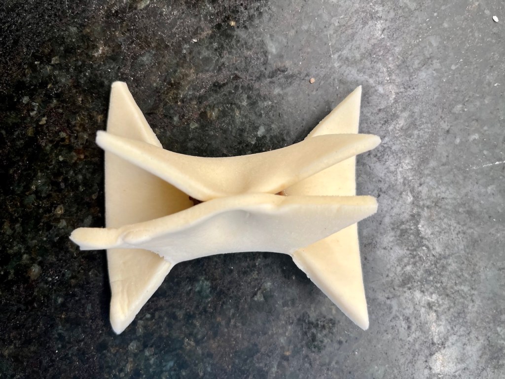 A top-down view of a uniquely shaped, white dough object resembling a bowtie, resting on a dark textured surface.