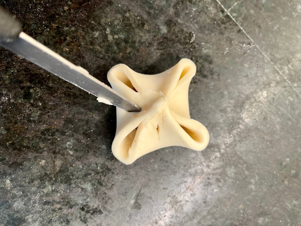 A close-up of a piece of dough shaped like a star with four pointed edges, resting on a granite surface, with a knife positioned nearby.