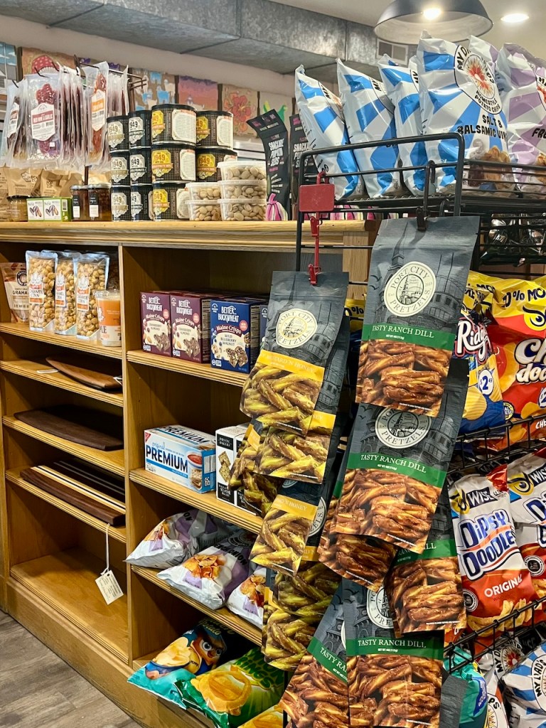 A colorful display of various snack foods, including pretzels, chips, candy, and jars of preserves, arranged on wooden shelves in a store. The scene showcases a variety of packaging designs and products.