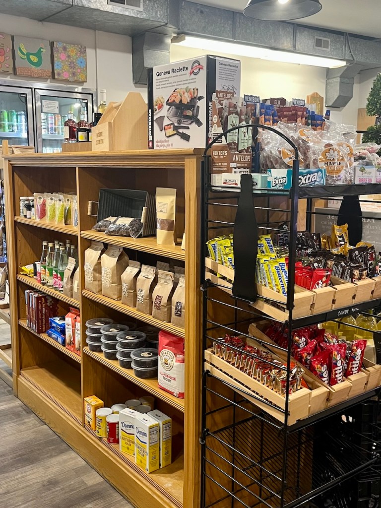 A wooden display shelf and a black metal rack in a store showcasing a variety of snacks, beverages, and groceries, including packaged meats, candy, sauces, and baking ingredients.