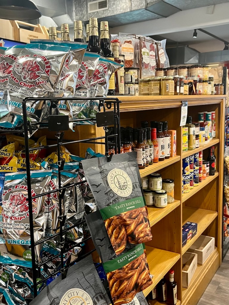 A display of snacks including various potato chips and pretzels on racks, alongside shelves filled with sauces and condiments in a grocery store.