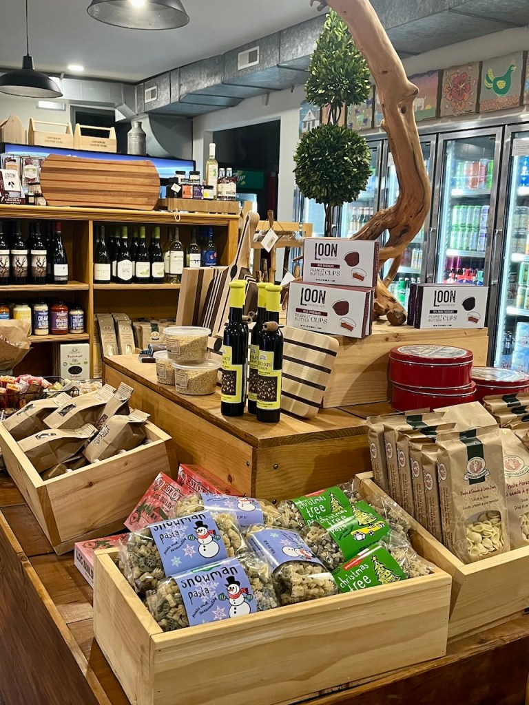 A cozy display of gourmet food items and gifts in a wooden shop interior, featuring various packaged snacks, bottles of oils, and festive pasta shapes.