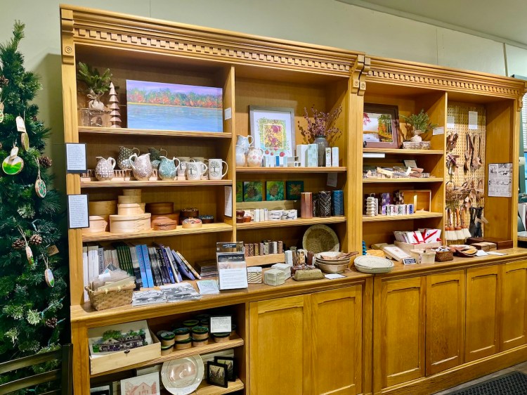 A wooden display shelf showcasing a variety of handcrafted items, including pottery, art, wooden bowls, and books, alongside a decorated Christmas tree in the corner.