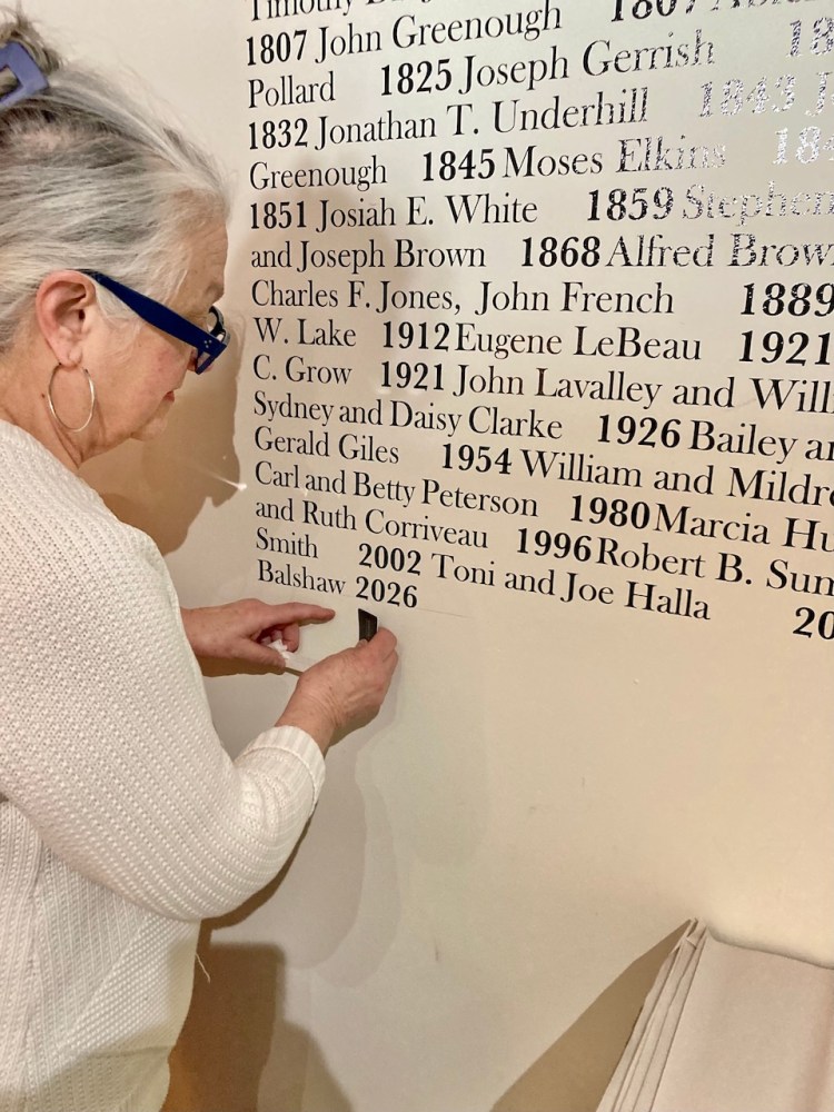A person with gray hair and glasses places a nameplate on a wall inscribed with names and years, indicating an ongoing historical or memorial project.