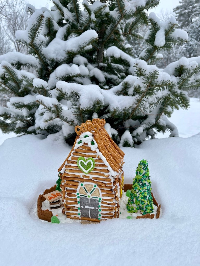 A gingerbread house decorated with icing and candy, surrounded by snow-covered ground and evergreen trees.