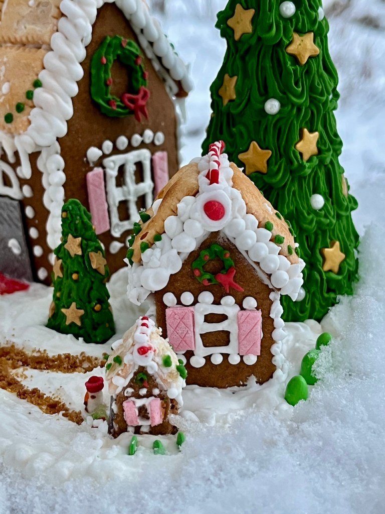 A festive gingerbread house decorated with icing, surrounded by a small gingerbread cottage and a green, textured Christmas tree, all set in a snowy landscape.