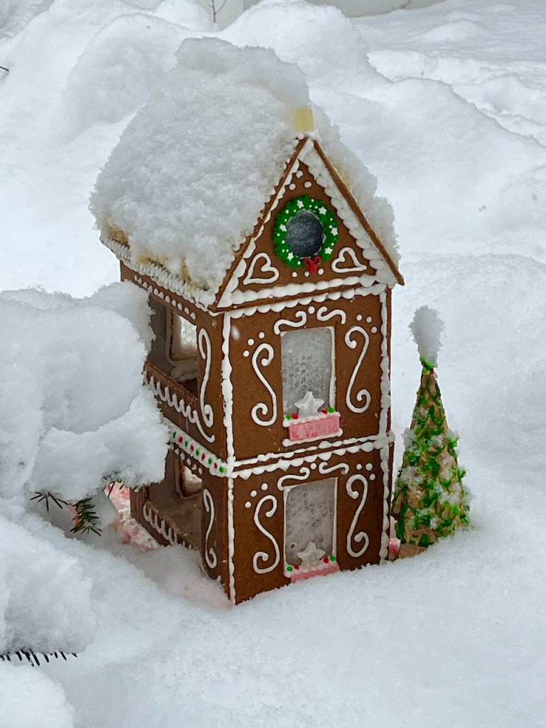 A decorated gingerbread house topped with snow, surrounded by white snow and featuring a small candy tree.