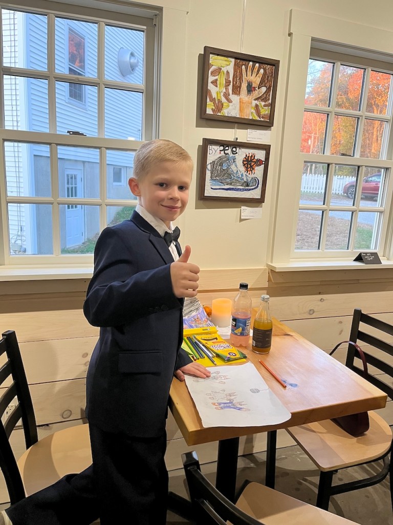 A young boy in a formal suit giving a thumbs up, standing at a table with drawings and art supplies, in a room with bright windows and artwork on the walls.